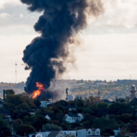 Large fire at an oil refinery seen from a distance. Only parts of the refinery visible behind the trees. Thick black smoke rises from the flames.