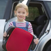 Portrait Of Girl Holding Booster Seat Standing Next To Car