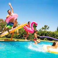 Group of age-diverse boys and girls, happy friends with swim floats jumping into swimming pool on the vacations