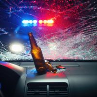A shattered windshield with a beer bottle, viewed from inside a car, illuminated by police lights in the background. This depicts the consequences of drunk driving.