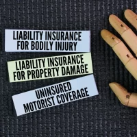 A wooden hand rests next to three cards on a textured black surface. The cards read: "Liability Insurance for Bodily Injury," "Liability Insurance for Property Damage," and "Uninsured Motorist Coverage."
