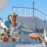 Two seafarers painting ship's crane while out at sea. Both wearing full PPE and fall protection harnesses to prevent fall and injury. Onboard safety culture concept.