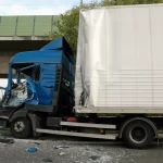 A blue truck with a crushed cab has collided with the rear of another truck, causing debris on the road. A person in a yellow safety vest is nearby.
