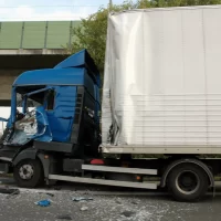 A blue truck with a crushed cab has collided with the rear of another truck, causing debris on the road. A person in a yellow safety vest is nearby. A blue truck with a crushed cab has collided with the rear of another truck, causing debris on the road. A person in a yellow safety vest is nearby.