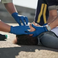 Two emergency responders tend to a person lying on the ground, wearing blue gloves. The scene conveys urgency and care in a medical situation.